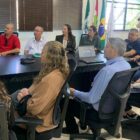 Doze pessoas em volta de uma mesa, entre homens e mulheres, olham para frente para acompanhar apresentação