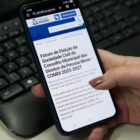 Mão de mulher segura celular perto de teclado. Na tela do aparelho, há informações sobre o fórum para eleger conselheiros para o Conselho do Idoso