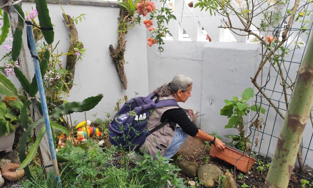 Mulher com colete e mochila está agachada em jardim virando um vaso que continha água