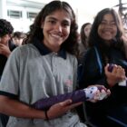 Duas alunas com uniforme seguram guarda-chuva e sorriem para câmera