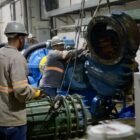 Homens com capacetes e equipamentos de segurança trabalham em grande equipamento na ETA Cubatão