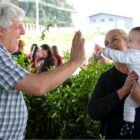 Homem grisalho brinca com bebê no colo de uma mulher, eles estão em uma feira perto de stand com plantas