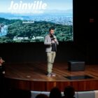 Homem branco segura microfone em apresentação em palco, com painel ao fundo com as palavras Joinville, o futuro do turismo