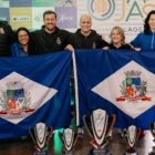 Sete pessoas, entre homens e mulheres, seguram duas bandeiras de Joinville, atrás de cinco troféus do Parajasc