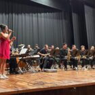Em palco, mulher com vestido vermelho segura microfone e canta. Ao seu lado, há uma orquestra com vários músicos tocando sentados