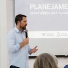 Secretário de educação, homem com cabelo e barba castanhos, segura microfone e fala para plateia em frente a um telão com a palavra "planejamento"