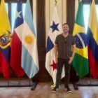 Prefeito Adriano Silva, um homem de meia idade, branco e com cabelo castanho, posa para foto sorrindo na frente de bandeiras de vários países