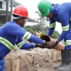 Dois homens uniformizados e com capacete manuseiam tubo usado na rede de esgoto