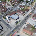 Imagem aérea com duas ruas principais e outras duas menores perpendiculares, há casas, comércios e carros trafegando