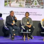Dois homens e uma mulher sentados em cadeiras em palco. Um deles segura o microfone e fala para plateia