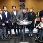 Homem em pé assina documento enquanto outros homens e mulheres ao lado assistem ao ato