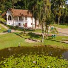 Casa branca ornamentada aparece ao fundo da imagem que também mostra um lago, gramado e vegetação