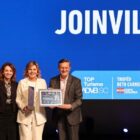 Quatro pessoas em pé no palco estão sorrindo e o telão no fundo mostra a palavra Joinville