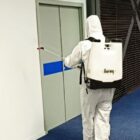 Pessoa usa equipamento de proteção individual branco e carrega pulverizador nas costas enquanto pulveriza produto em parede