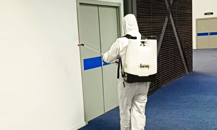 Pessoa usa equipamento de proteção individual branco e carrega pulverizador nas costas enquanto pulveriza produto em parede