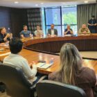 Dezenas de pessoas, homens e mulheres de várias idades, estão sentados ao redor de uma mesa com formato arredondado em uma sala