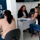 Duas mulheres sentadas estão em frente a computadores enquanto atendem outras duas mulheres sentadas
