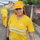 Mulher vestida com boné e camisa amarela segura vassoura na mão e olha para a câmera