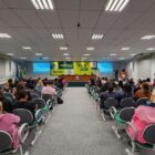 Amplo auditório com dezenas de pessoas sentadas de frente para um palco