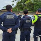 Dois homens vestidos com uniformes azuis da Guarda Municipal, um homem vestido com uniforme azul do Detrans e outro homem vestido com uniforme marrom da Polícia Militar estão em pé às margens de uma rua