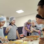 Crianças usando toucas descartáveis manuseiam alimentos sobre uma mesa incentivadas por duas mulheres