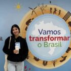 Garota com cabelos castanhos posa para foto e sorri na frente de banner com a frase "vamos transformar o Brasil"