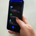 Mão segura celular com app Joinville Fácil aberto, mostrando abas como tábua de marés e câmeras em tempo real