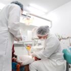 Duas pessoas de roupa branca e toca descartável atendem criança em consultório odontológico