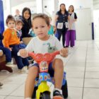 Menino sentado em motoquinha olha para a câmera enquanto crianças e adultos observam ao seu redor