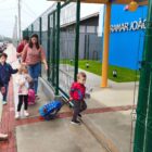 Imagem mostra portão de CEI aberto enquanto criança vestida de blusa vermelha entra com a mochila. Adultos e outras crianças também se aproximam do portão