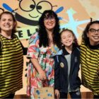 Menina sorri em pé ao lado da sua professora e de duas mulheres vestidas de abelha, com roupas listradas em preto e amarelo