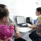 Mulher sentada com criança no colo em consultório é atendida por outra mulher sentada na frente de computador