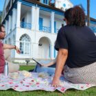 Três pessoas sentadas em uma toalha no gramado na frente de um museu, uma construção antiga