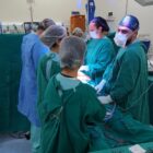 Equipe médica com vestimenta verde está em pé em sala de cirurgia e algumas pessoas portam instrumentos cirúrgicos