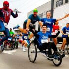 Homem em triciclo é conduzido por outro homem e mulher, todos olham para a câmera e sorriem, ao lado de homem vestido de Homem Aranha que dá um salto