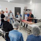 Pessoas usando toucas descartáveis se reúnem em círculo observando homem de avental e carne exposta em uma bancada de inox