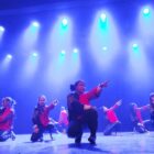 Dançarinos com figurino estão no palco, parcialmente ajoelhados, e apontando em diferentes direções durante apresentação de dança