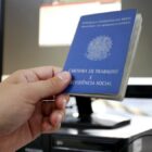 Mão segura uma carteira de trabalho, documento retangular azul, em frente a um computador