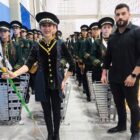 Alunos da fanfarra estão posicionados em pé, em três filas, vestindo rupas e chapéus e portando instrumentos