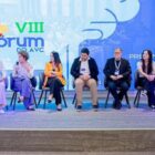 Oito homens e mulheres estão sentados em cadeiras em palco com painel que mostra o termo VII Fórum AVC