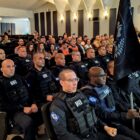 Homens uniformizados sentados em amplo auditório, um deles segura uma bandeira