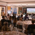 Homem em pé fala para dezenas de pessoas sentadas em sala