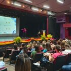 Mulher está no palco com microfone na mão e aponta para telão enquanto pessoas sentadas assistem à apresentação