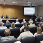 Homem com microfone fala na frente de sala com dezenas de pessoas sentadas