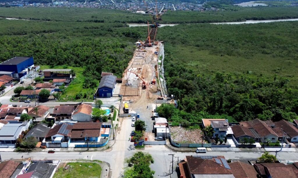 Imagem aérea mostra casas, ruas, área de vegetação, rio e canteiro de obras onde está sendo construída a Ponte Joinville