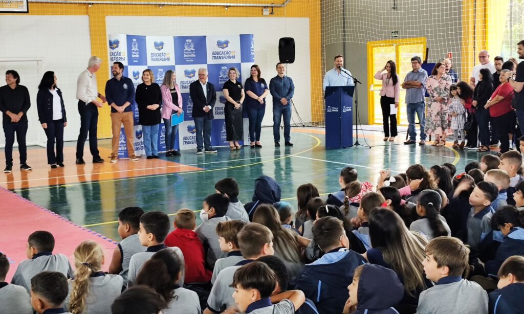 Alunos estão sentados na quadra enquanto lideranças políticas, homens e mulheres, estão à frente em pé