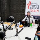 Em um estúdio de rádio, um homem entrevista outro e banner mostra o termo Rádio Joinville Cultural