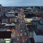 Imagem aérea de avenida com três pistas para carros, está de noite, há outros carros estacionados, comércios e casas