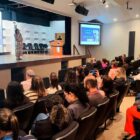 Diversas pessoas estão sentadas à frente de um palco onde mulher com microfone está falando
