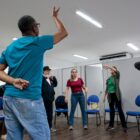 Em uma sala, várias pessoas em pé fazem gestos diferentes em aula de teatro para idosos